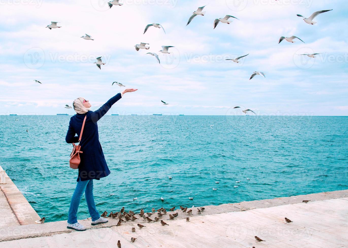 blonde woman feeding seagulls in cloudy autumn day on the sea coast photo