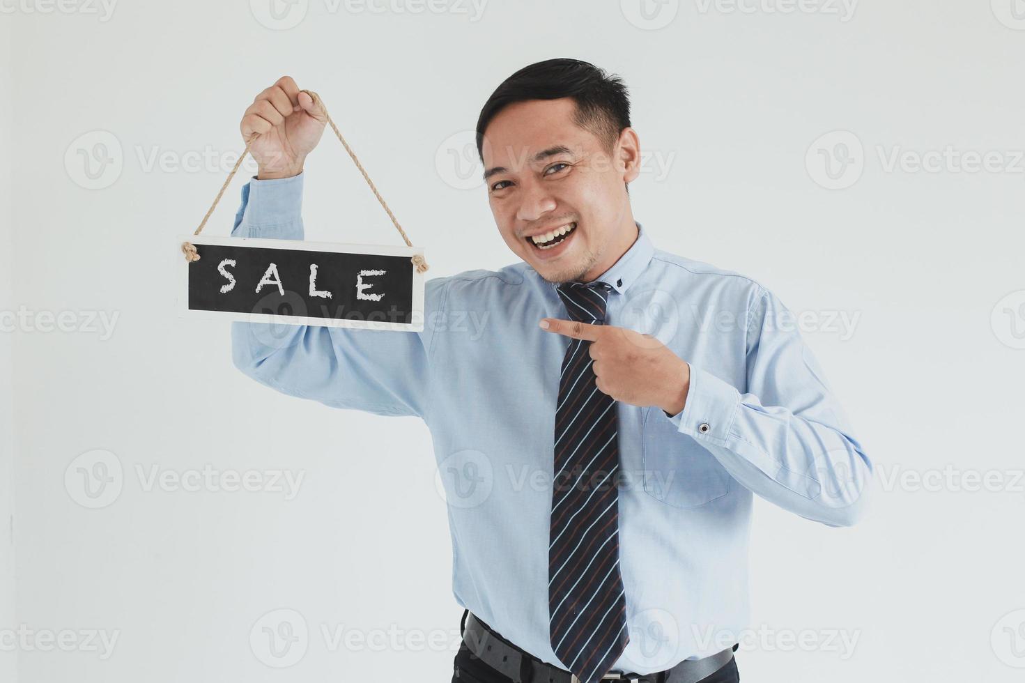 Vendedor sonriente vestido con camisa azul y corbata posando con señalar y llevar cartel de venta sobre fondo blanco. foto