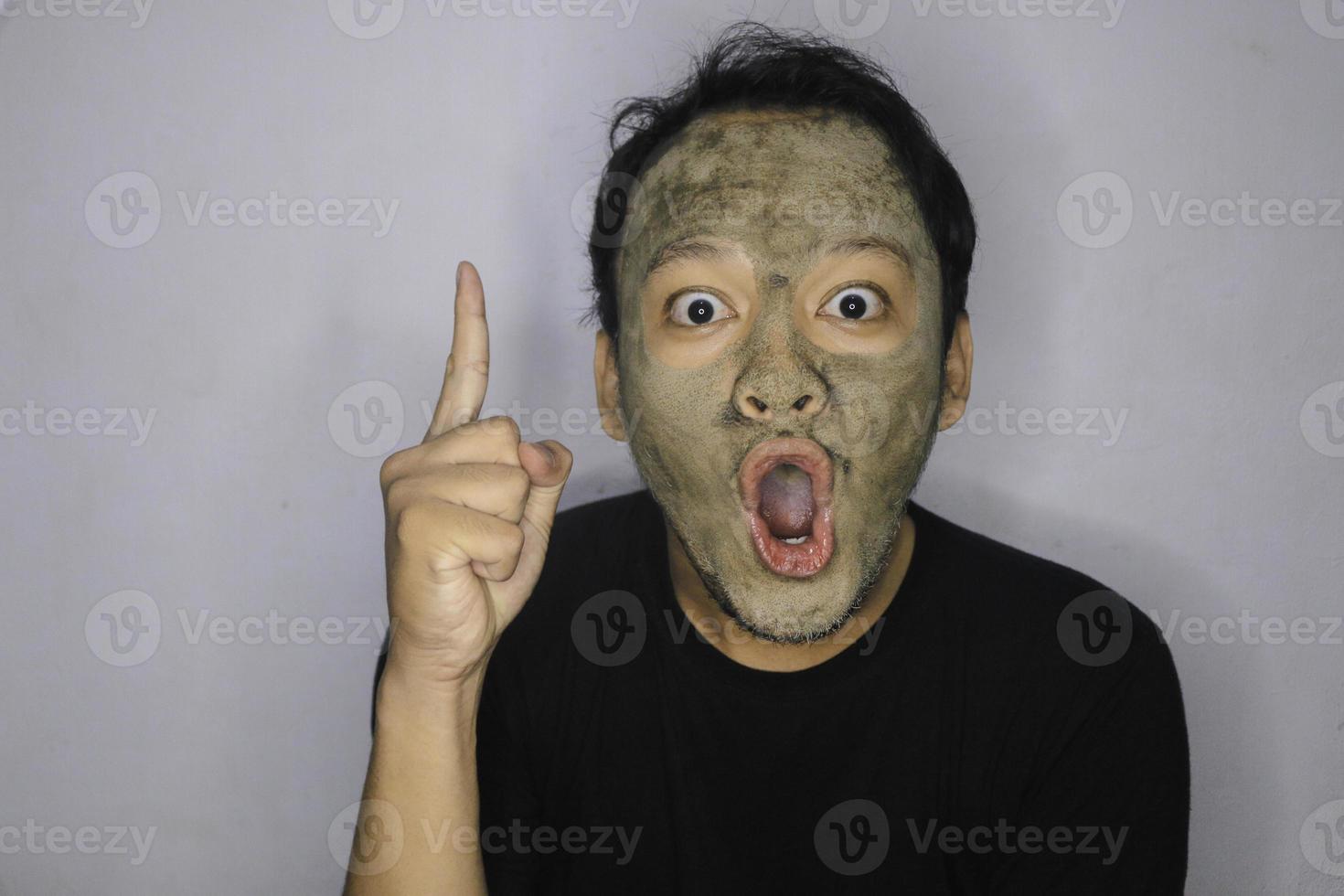sorprende al hombre asiático que señala arriba y mira a la cámara cuando usa una máscara facial de belleza foto