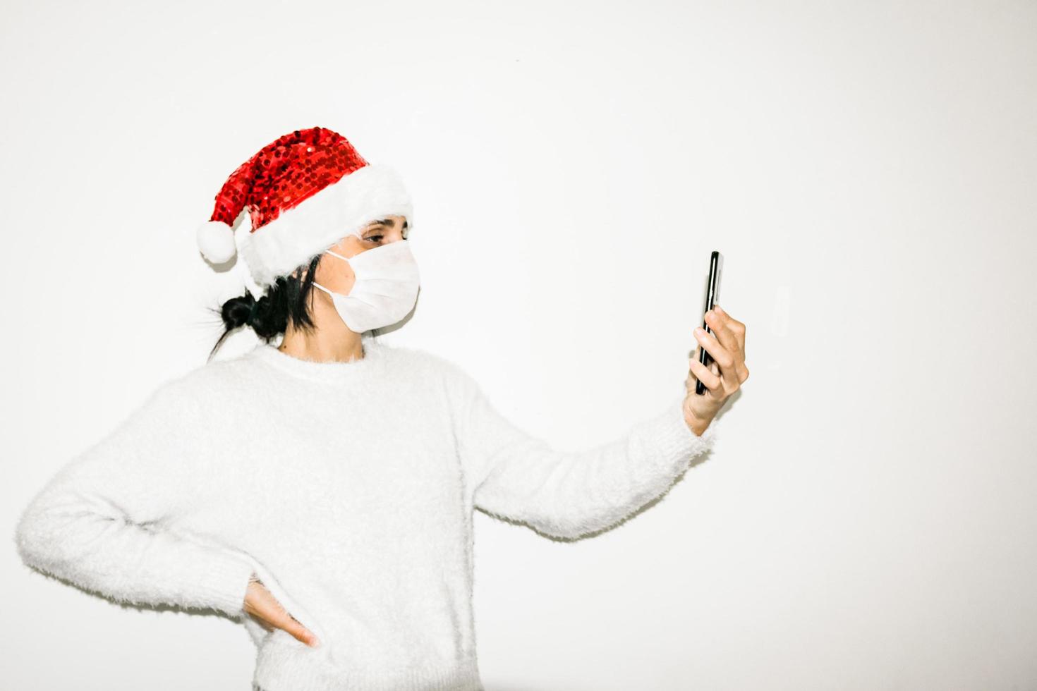 Young woman with facemask on holds cellphone high up isolated in white background. Concept of facetime and cummunication during holidays on pandemic. photo
