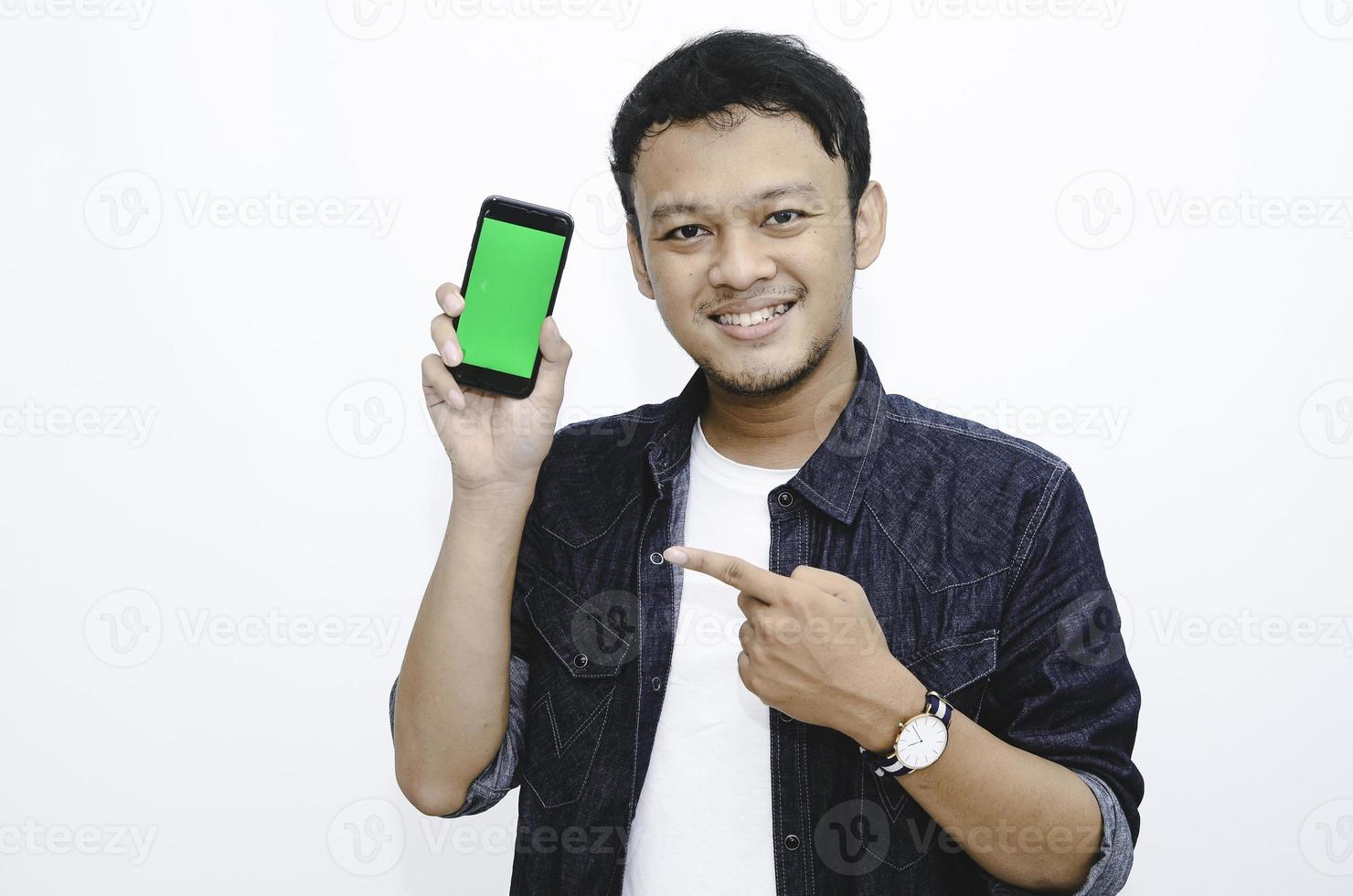 Young Asian man is standing and smiling pointing on green screen on smartphone screen on white background. photo