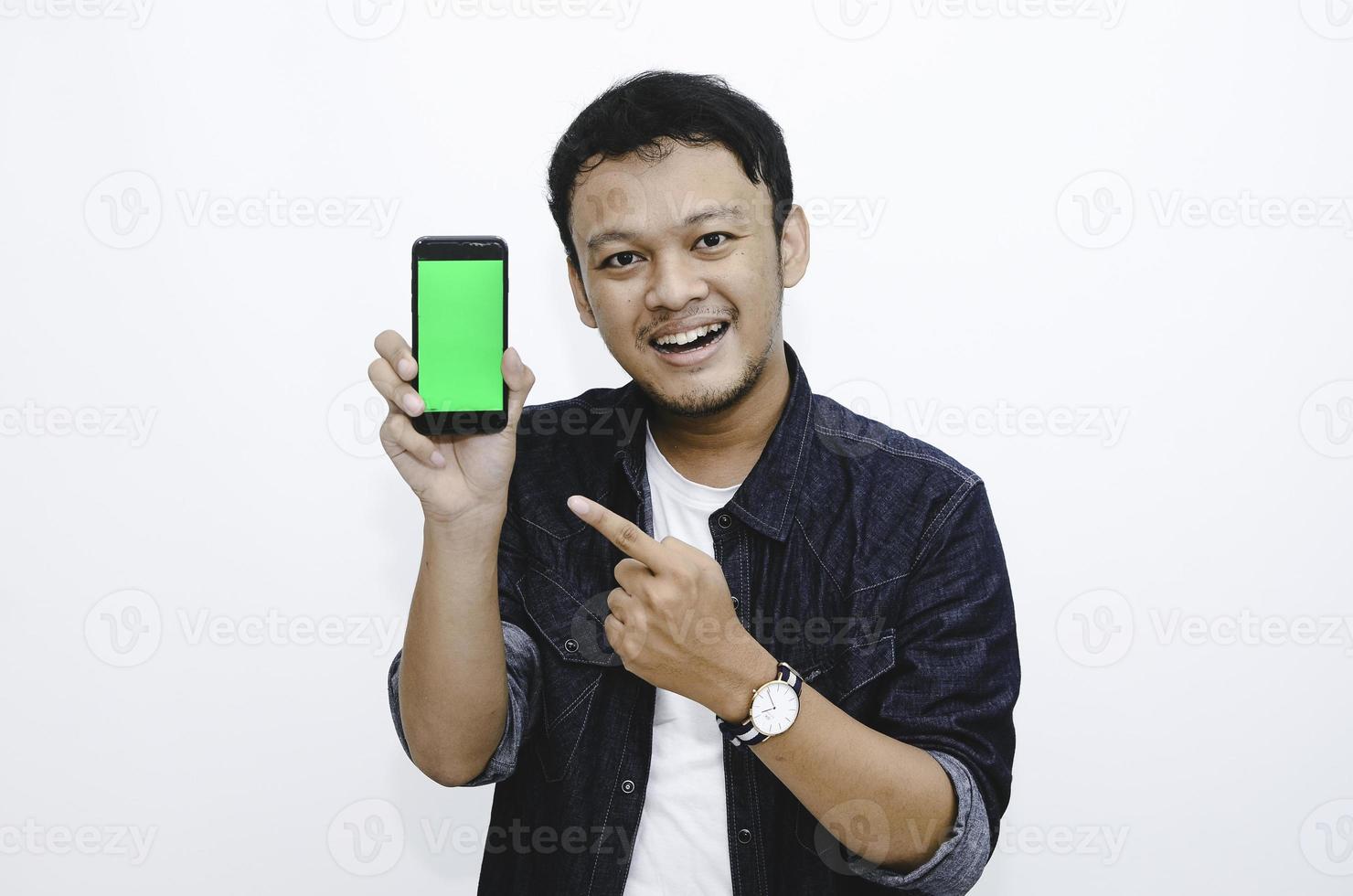 Young Asian man is standing and smiling pointing on green screen on smartphone screen on white background. photo