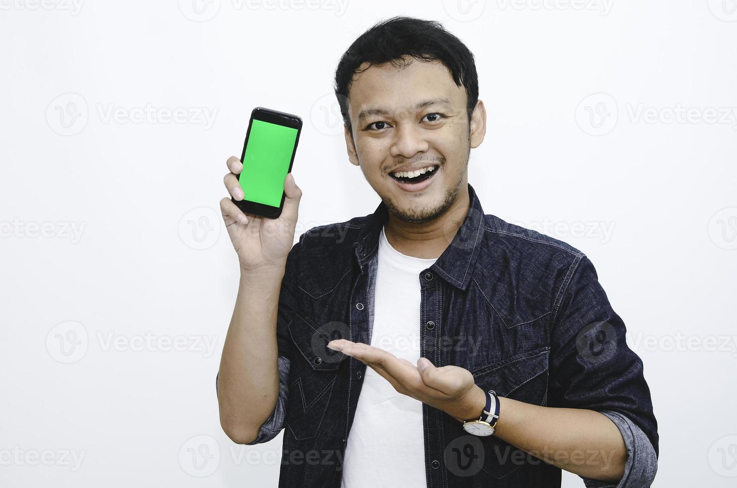 Young Asian man is standing and smiling pointing on green screen on smartphone screen on white background. photo