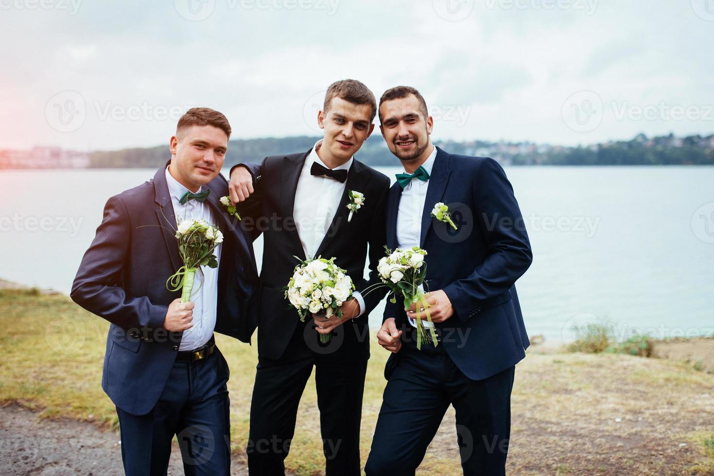 Novio guapo sonriente confiado en traje negro con dos padrinos de boda foto