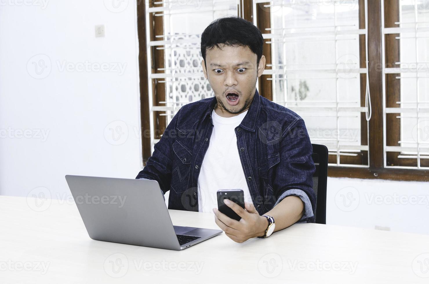 Young Asian man shocked and wow what he see in the laptop when in call on smartphone photo