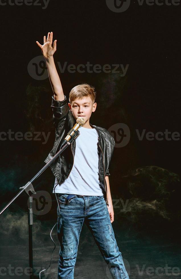 retrato de adolescente caucásico con camiseta blanca, jeans azules y chaqueta de cuero con micrófono cantando sobre fondo oscuro. concepto de hobby y gloria foto