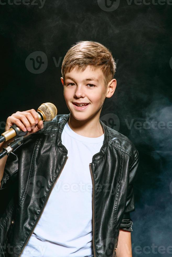 retrato de adolescente caucásico con camiseta blanca, jeans azules y chaqueta de cuero con micrófono cantando sobre fondo oscuro. concepto de hobby y gloria foto