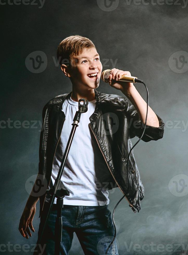 Portrait of caucasian teenager in white t-shirt, blue jeans and leather jacket with microphone singing on dark background. Hobby and glory concept photo