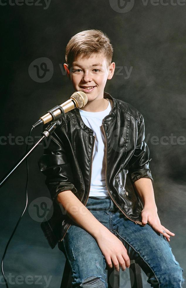 retrato de adolescente caucásico con camiseta blanca, jeans azules y chaqueta de cuero con micrófono cantando sobre fondo oscuro. concepto de hobby y gloria foto