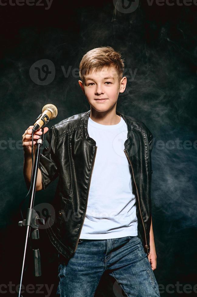 Portrait of caucasian teenager in white t-shirt, blue jeans and leather jacket with microphone singing on dark background. Hobby and glory concept photo