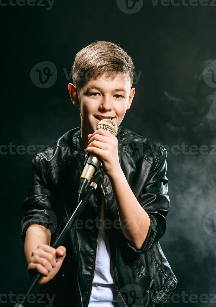 Portrait of caucasian teenager in white t-shirt, blue jeans and leather jacket with microphone singing on dark background. Hobby and glory concept photo