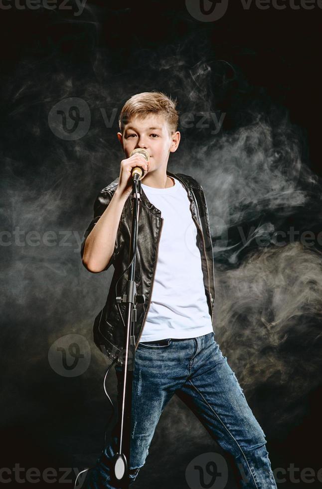 Portrait of caucasian teenager in white t-shirt, blue jeans and leather jacket with microphone singing on dark background. Hobby and glory concept photo