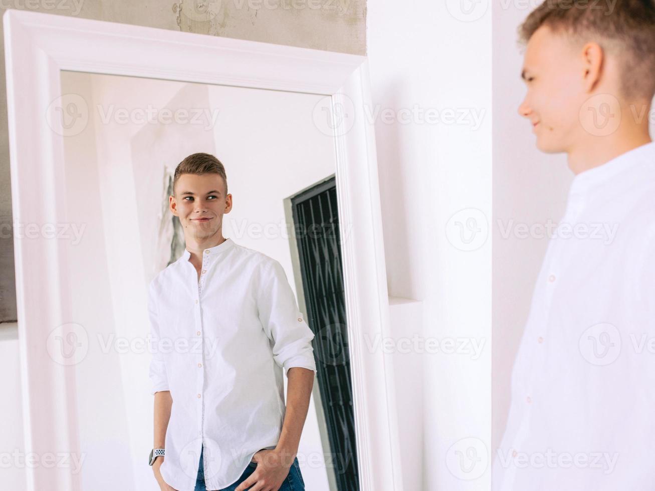 handsome guy in white shirt and blue jeans standing by the mirror indoor. Fashion, style concept photo