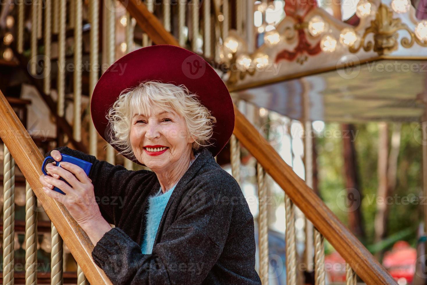 senior stylish woman in dark grey coat, hat and with grey hair standing by the carousel smiling, drinking coffee and enjoying life. Travel, fun, pension, happiness, seasonal concept photo