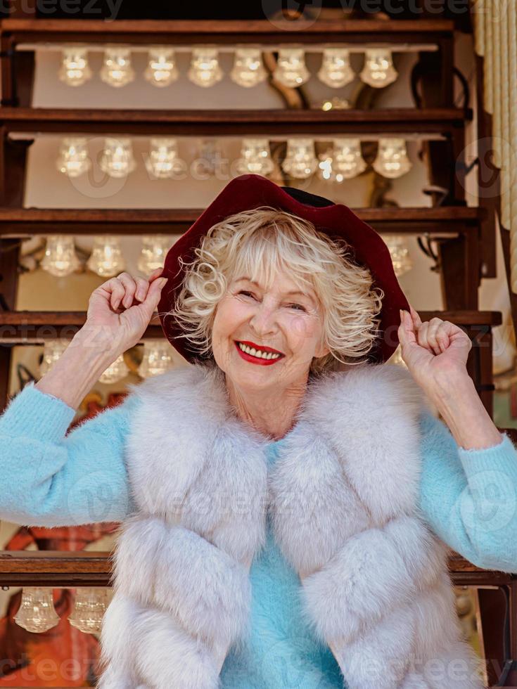 senior stylish woman in fur coat and with gray hair sitting on carousel holding purple hat. Travel, fun, fashion, happiness, seasonal concept photo