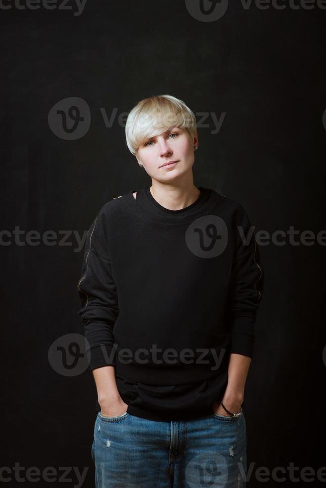 Portrait of stylish blonde short hair young woman in black sweatshirt and jeans on dark background photo