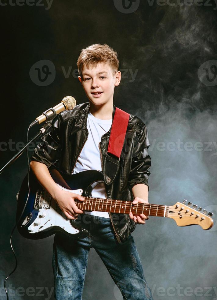 retrato de adolescente caucásico con camiseta blanca, jeans azules y chaqueta de cuero con micrófono y guitarra cantando en un fondo oscuro. concepto de hobby y gloria foto