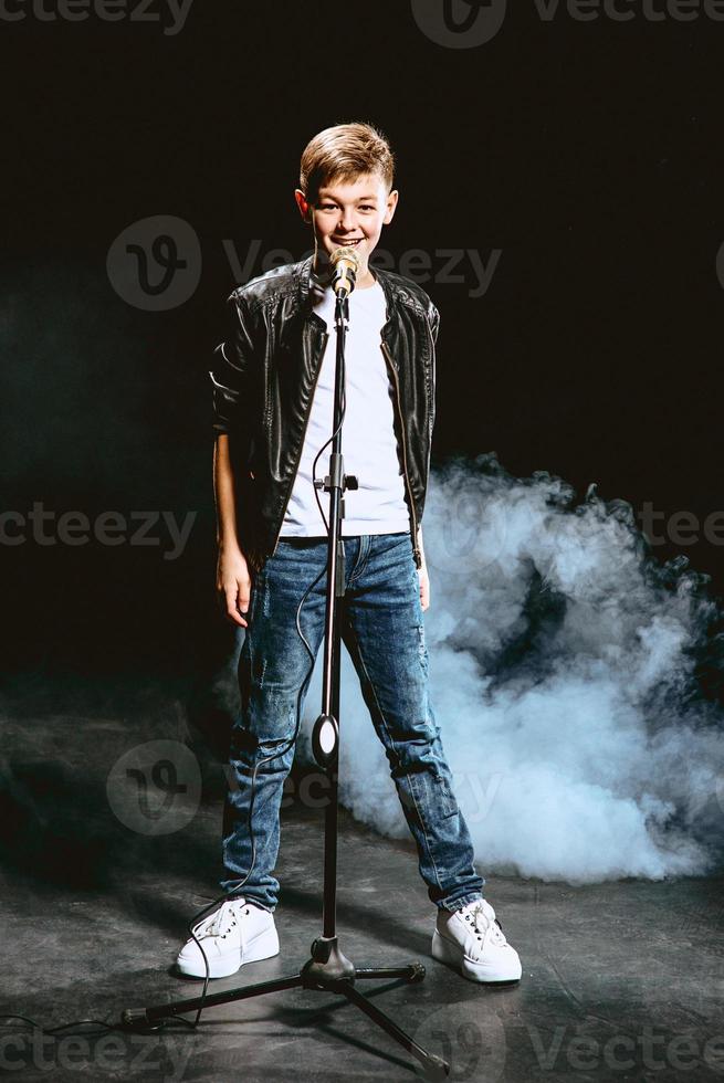 Portrait of caucasian teenager in white t-shirt, blue jeans and leather jacket with microphone singing on dark background. Hobby and glory concept photo