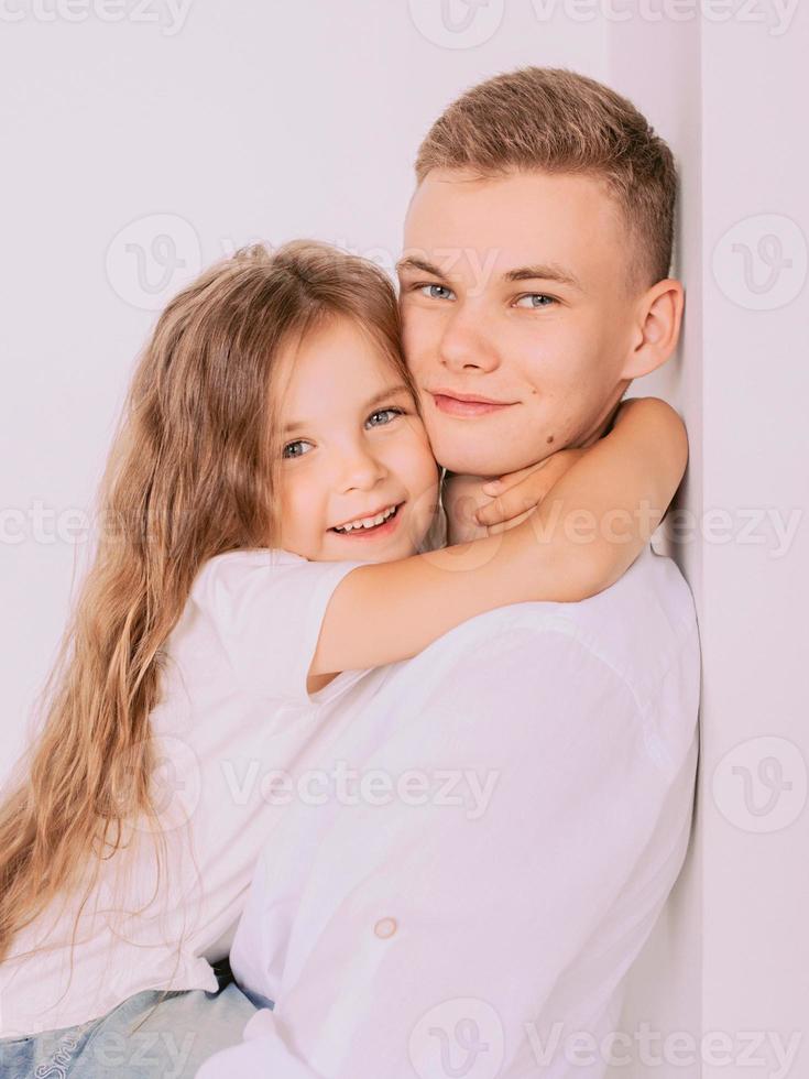 caucasian happy adorable siblings - teenager boy brother and little girl sister cuddling on white background indoor. Family, relationships, relatives, love, support concept photo