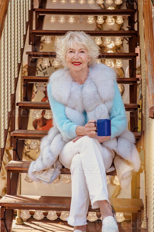senior stylish woman in fur coat and with grey hair sitting on carousel drinking tea and enjoying life. Travel, fun, happiness, seasonal concept photo