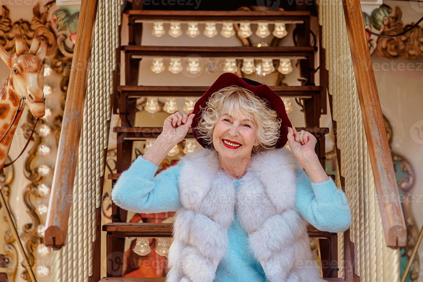 senior stylish woman in fur coat and with gray hair sitting on carousel holding purple hat. Travel, fun, fashion, happiness, seasonal concept photo