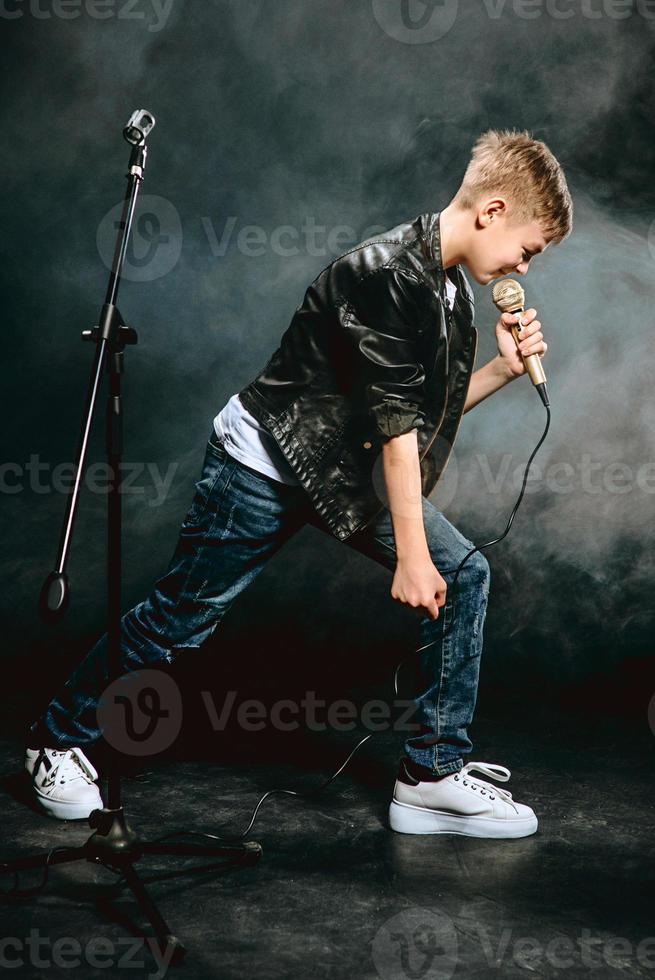 Portrait of caucasian teenager in white t-shirt, blue jeans and leather jacket with microphone singing on dark background. Hobby and glory concept photo