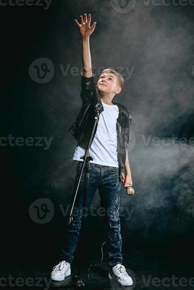 retrato de adolescente caucásico con camiseta blanca, jeans azules y chaqueta de cuero con micrófono cantando sobre fondo oscuro. concepto de hobby y gloria foto