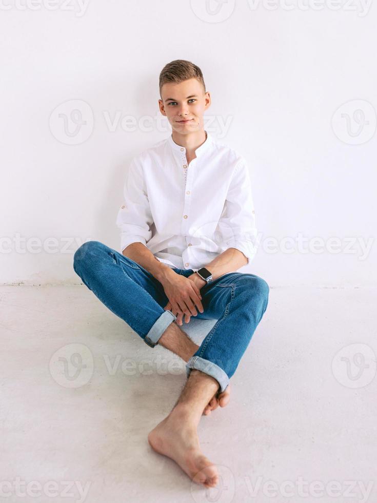 chico guapo con camisa blanca y jeans azules sentado en un piso interior. foto