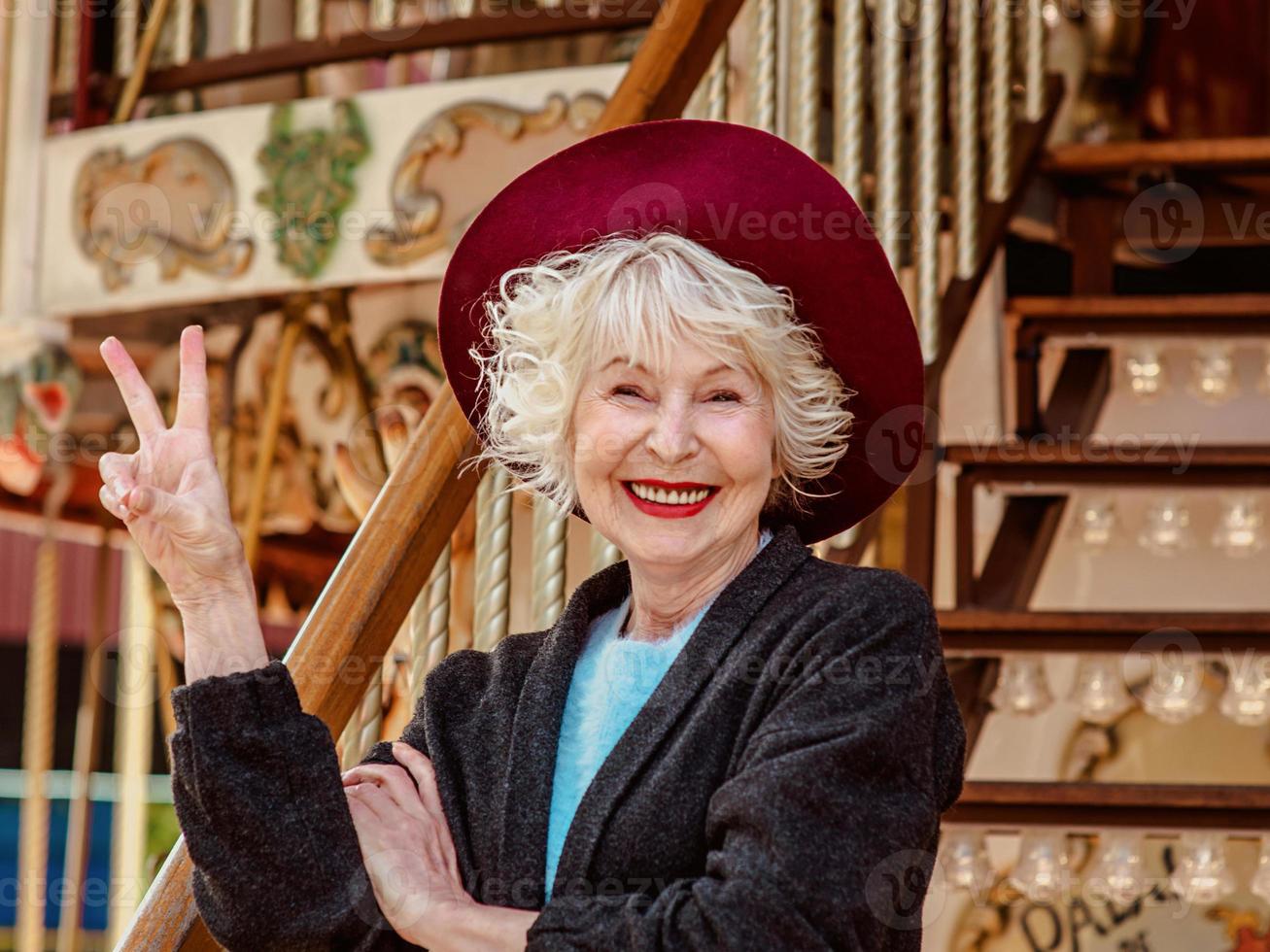 senior stylish woman in dark gray coat, hat and with grey hair standing by the carousel smiling and enjoying life. Travel, fun, pension, happiness, seasonal concept photo