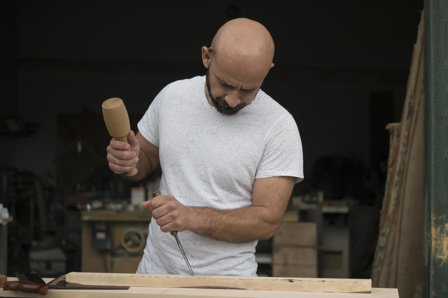 white bald carpenter with beard in white t-shirt works as a chisel and a mallet photo
