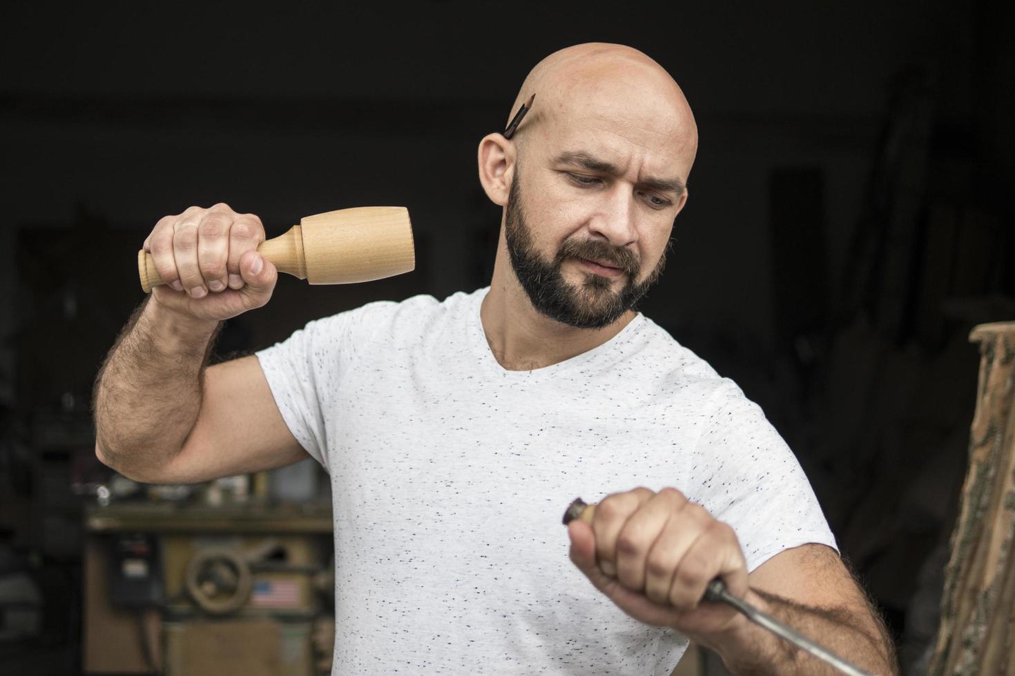 white bald carpenter with beard in white t-shirt works as a chisel and a mallet photo