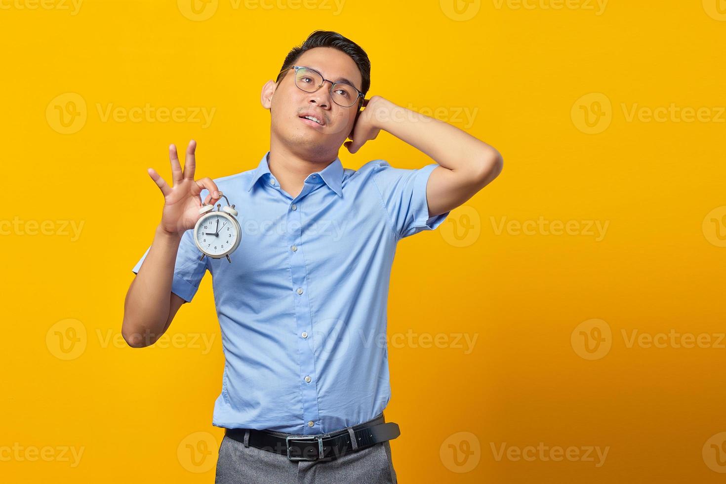 Pensive handsome Asian young man in glasses showing alarm clock and looking up isolated on yellow background. Time management lifestyle concept photo