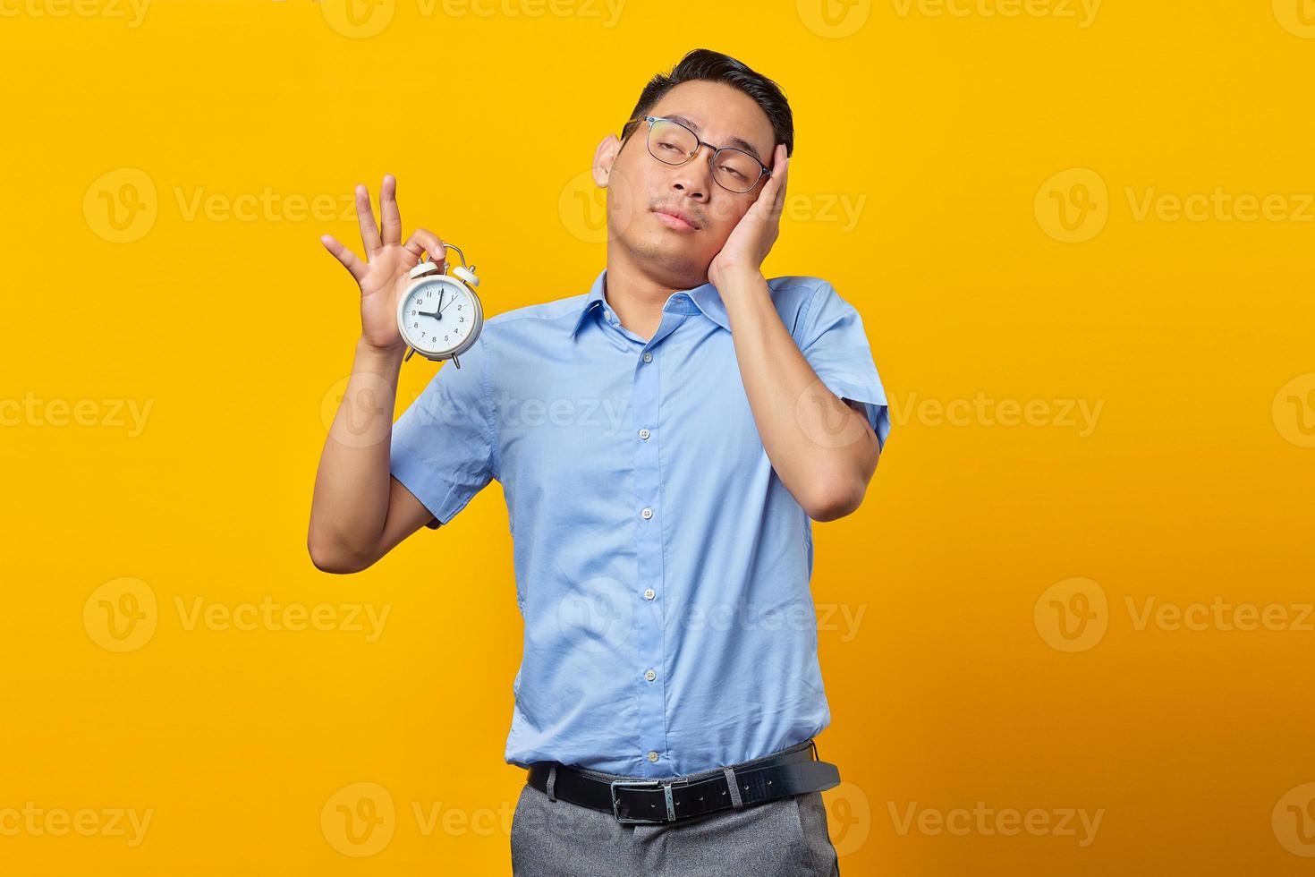 Portrait of handsome asian young man showing alarm clock feeling sleepy with closed eyes isolated over yellow background. Time management lifestyle concept photo