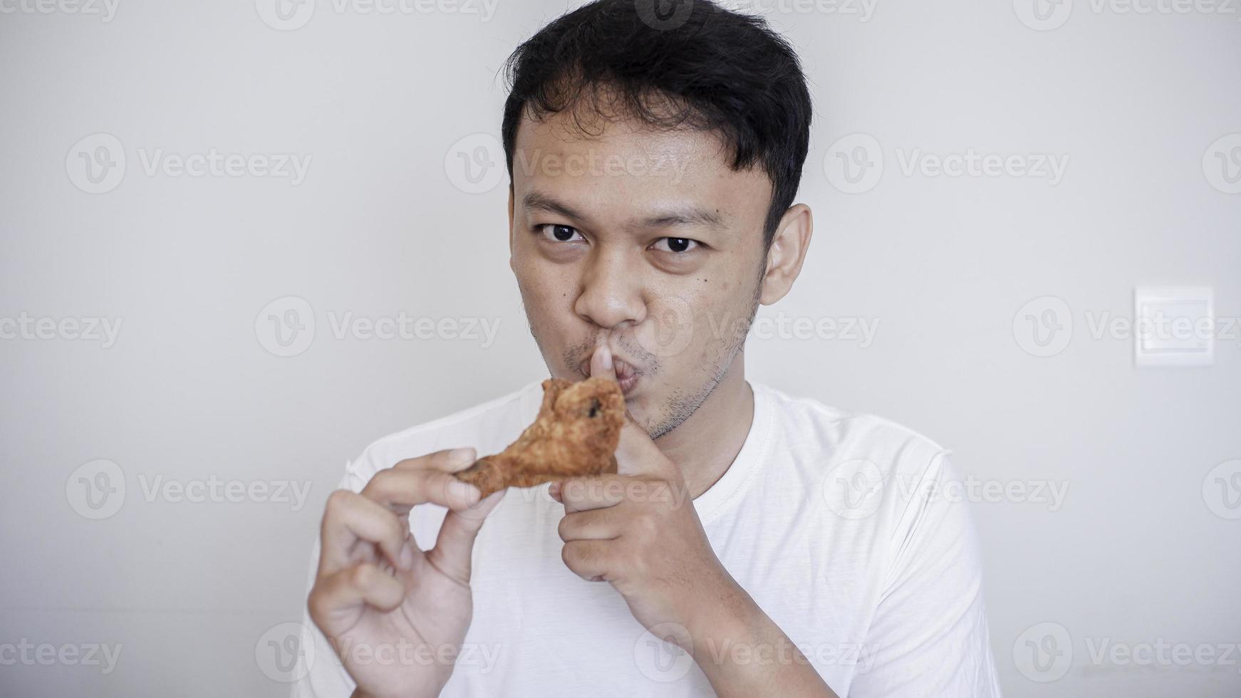 Young Asian man is eating fried chicken wear white shirt with silent hand gesture. photo