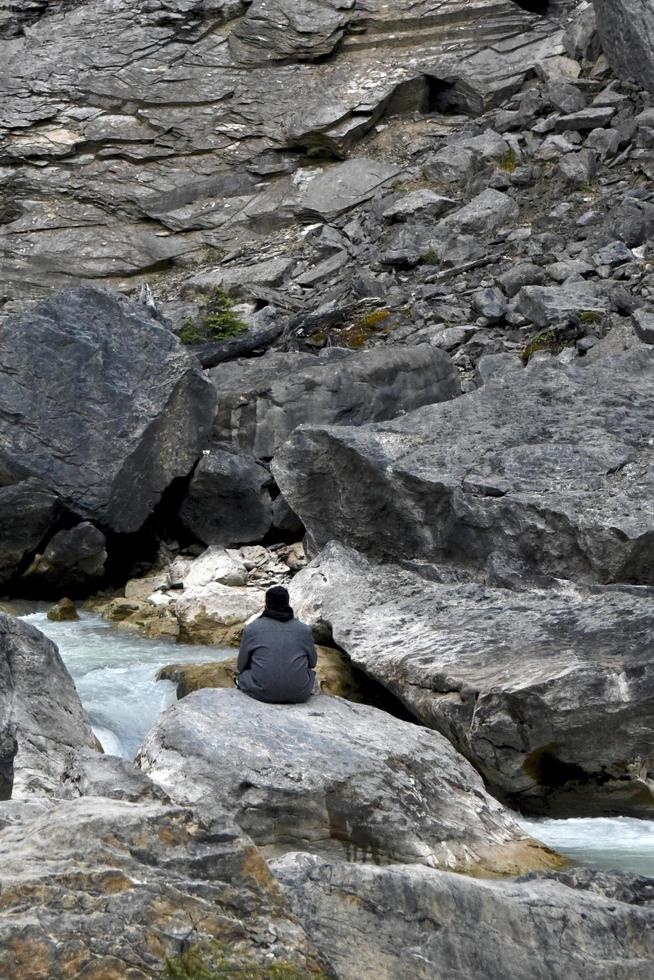 a man is seen from behind as he sits watching a rushing mountain stream photo