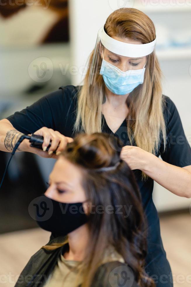 peluquero, protegido por una máscara, peinando el cabello de su cliente con una plancha de pelo en un salón. foto