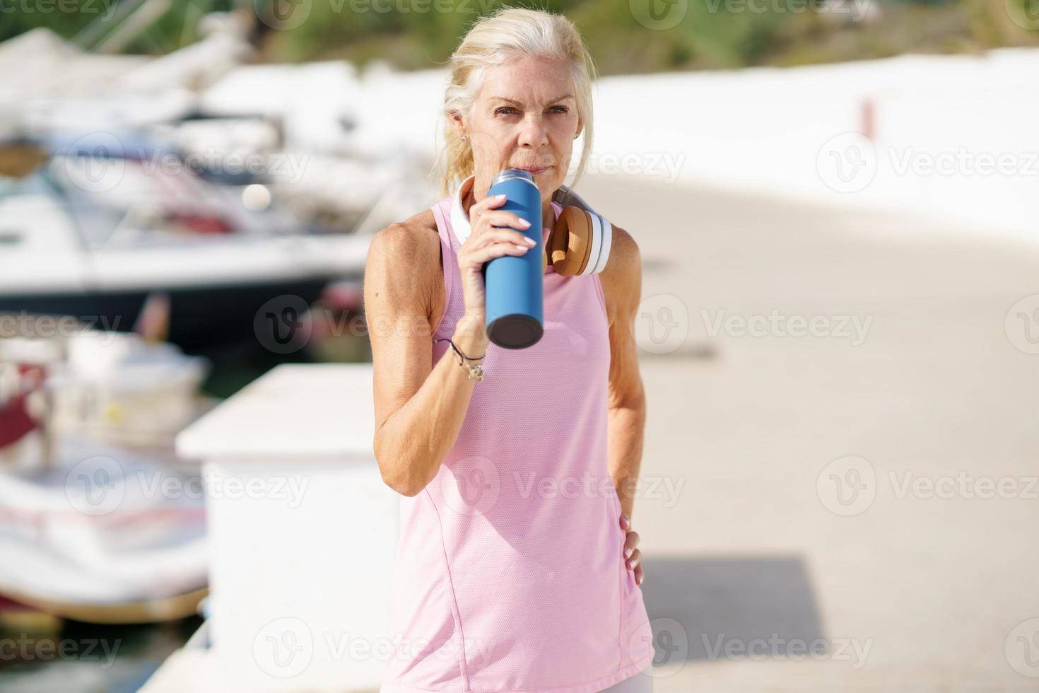 Mature sportswoman in fitness clothing drinking water from a metal fitness bottle outdoors. photo
