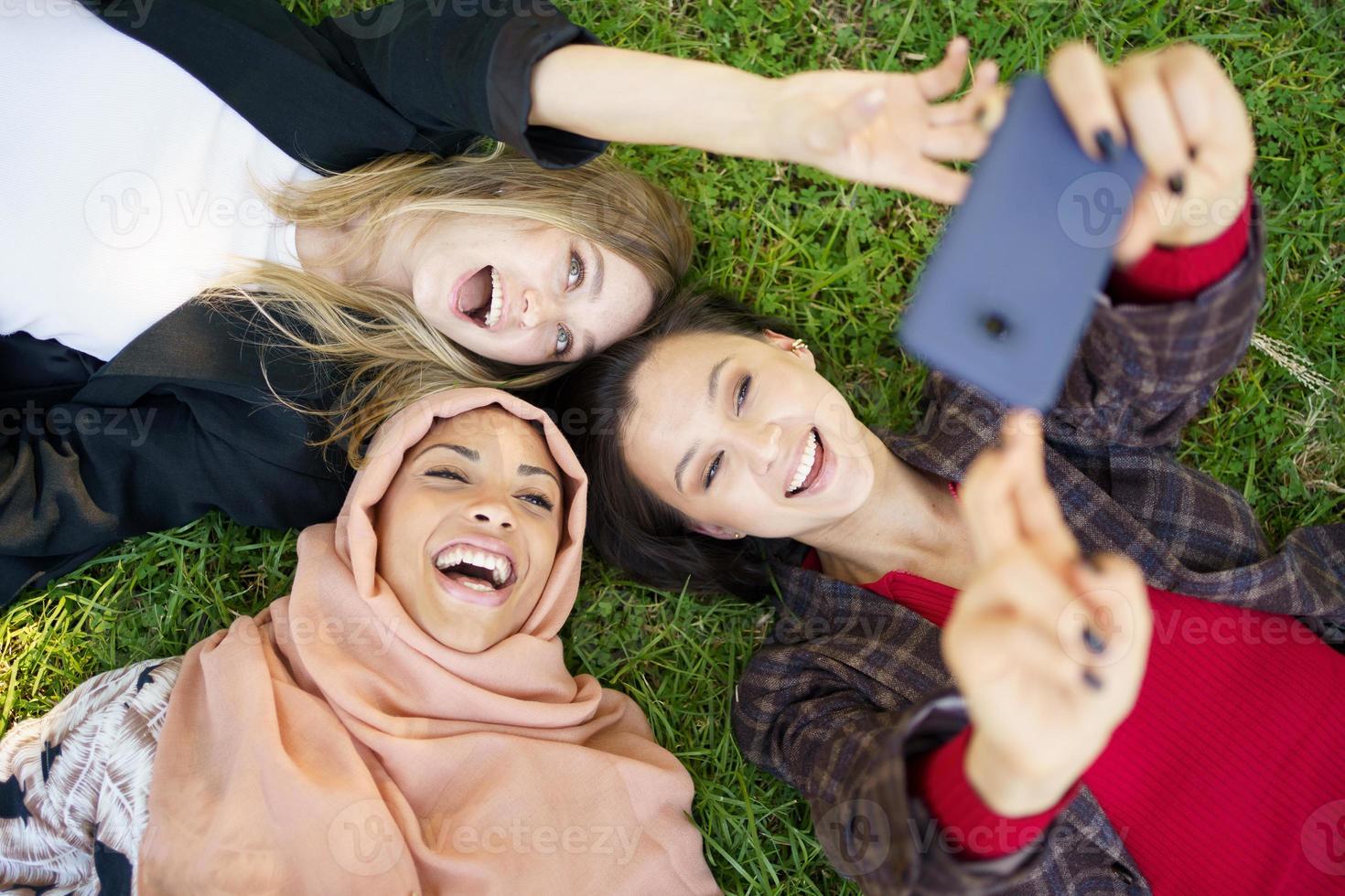 alegres y diversas novias riéndose y tomándose selfie tumbadas en el césped en el parque foto