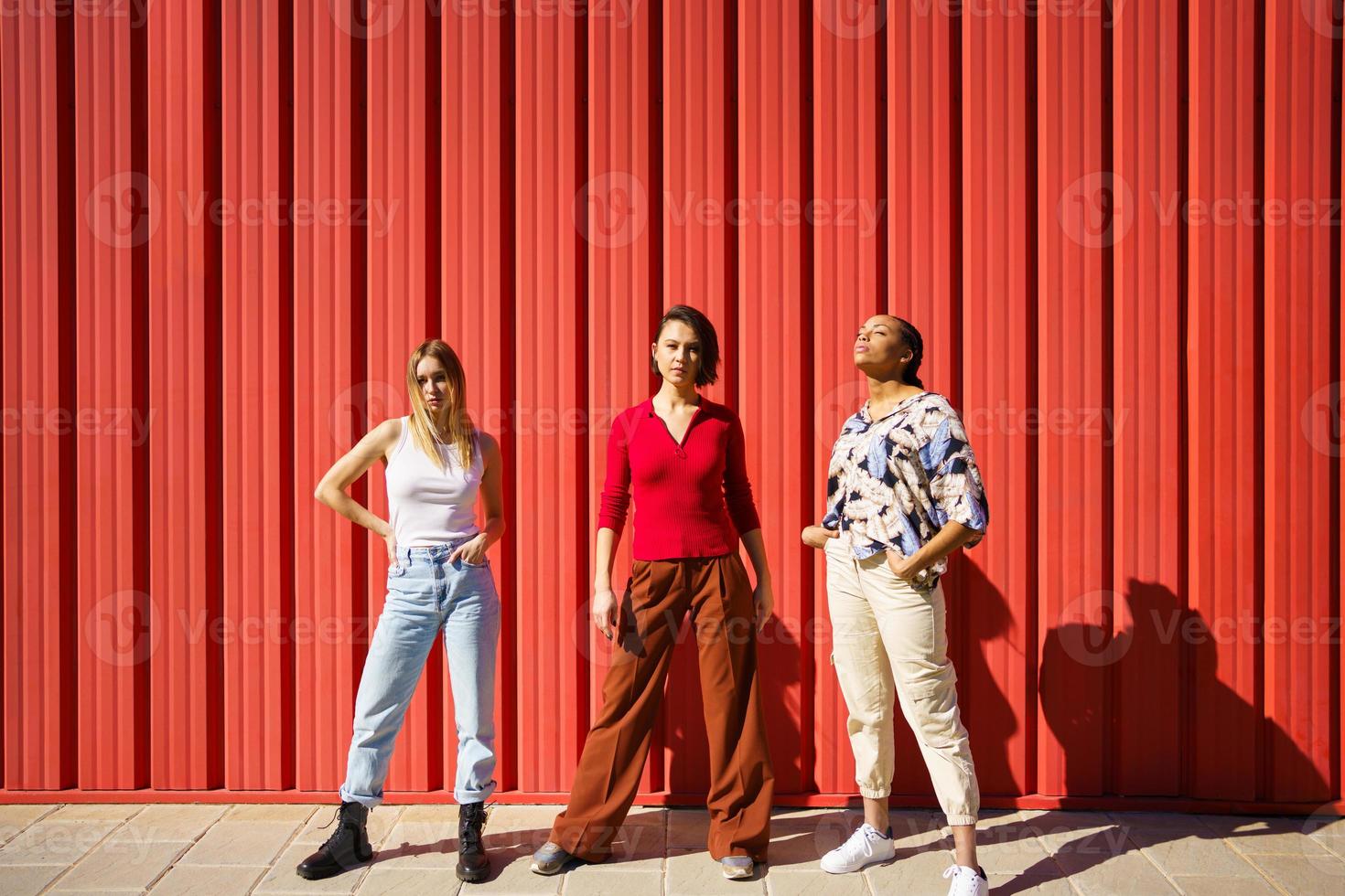 Stylish young diverse ladies standing against red fence on street photo