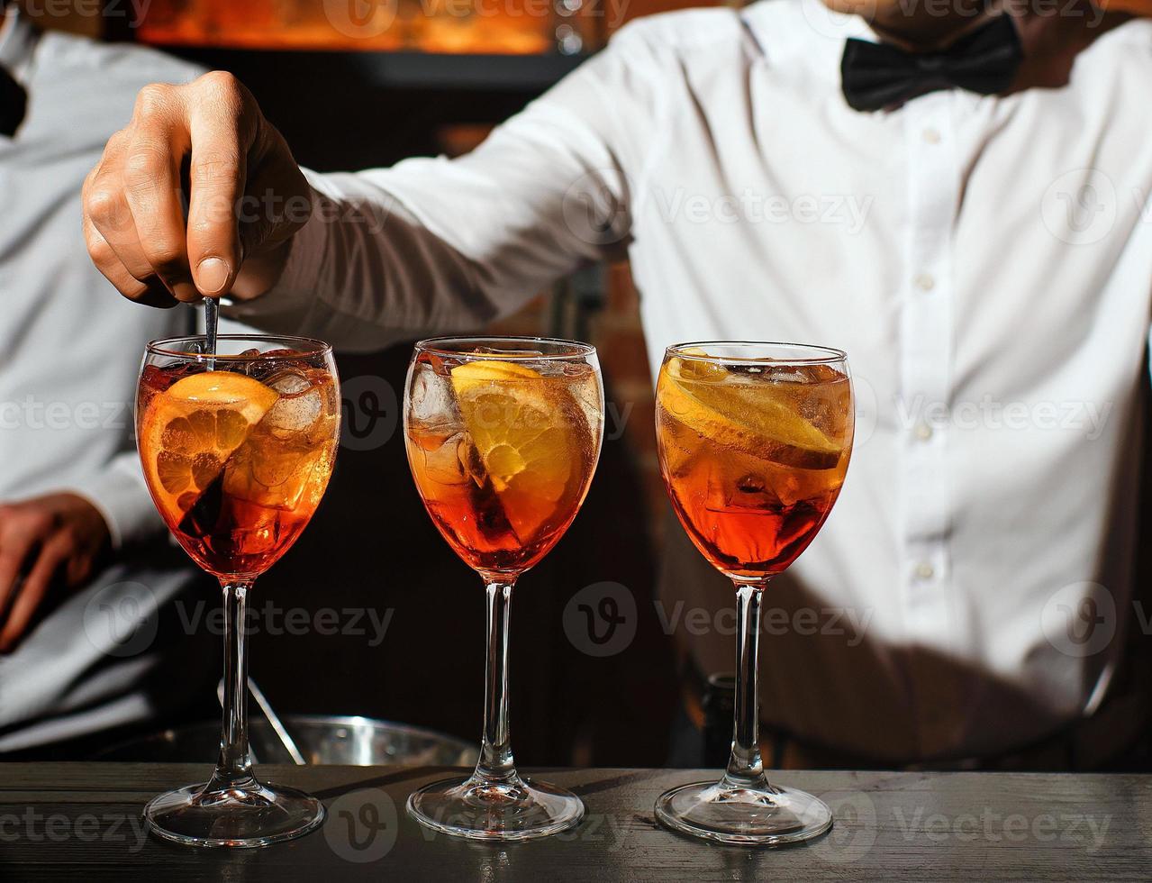 three glass glasses with an aperol spritz cocktail are on bar. bartender in white shirt, bow tie is stirring one of aperitifs. concept - people, drinks, entertainment, recreation. free space for text. photo