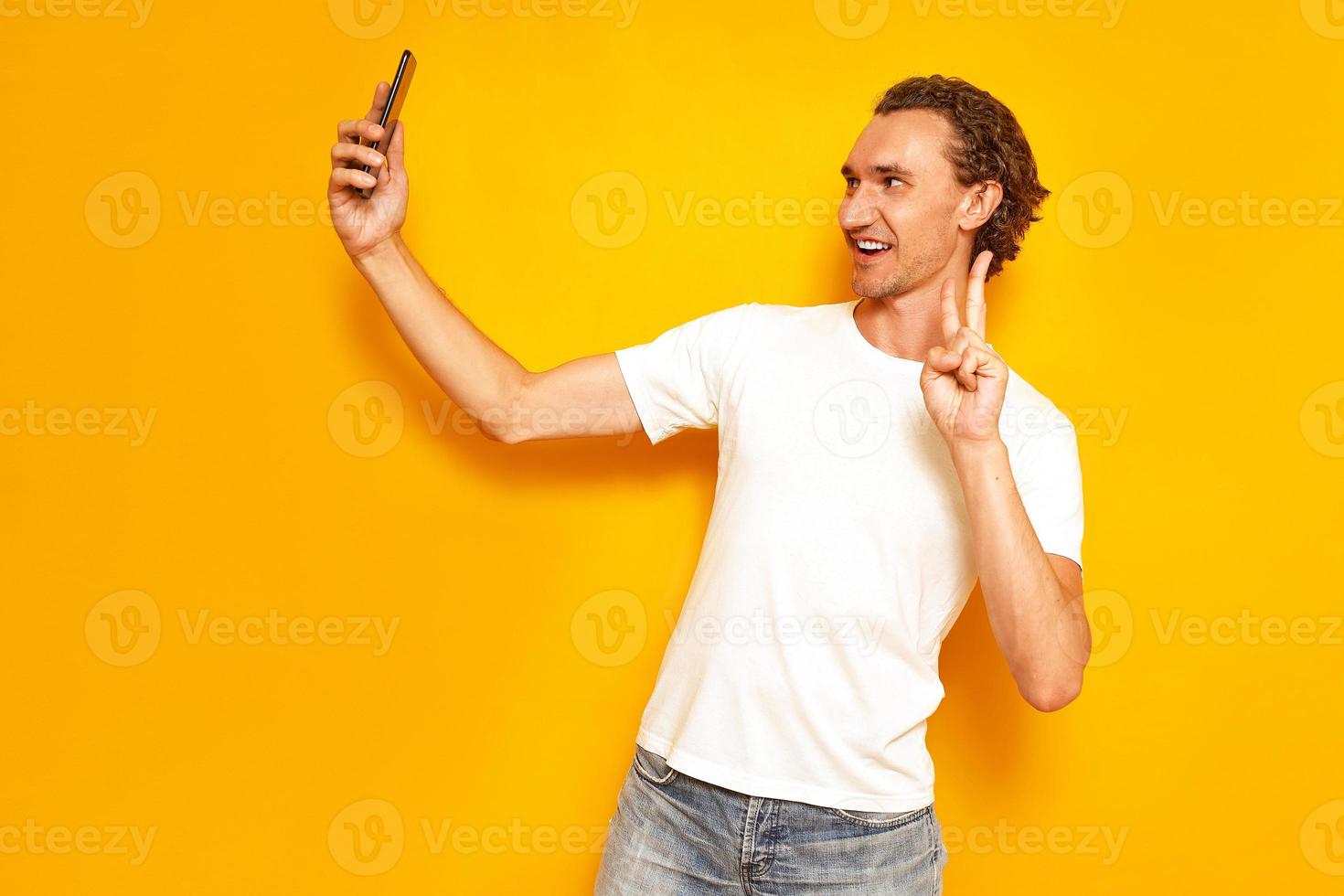 smiling man takes selfie on phone showing V sign of peace victory with hand on camera dressed in casual clothes. isolated on studio yellow background. concept - people, technology, video communication photo