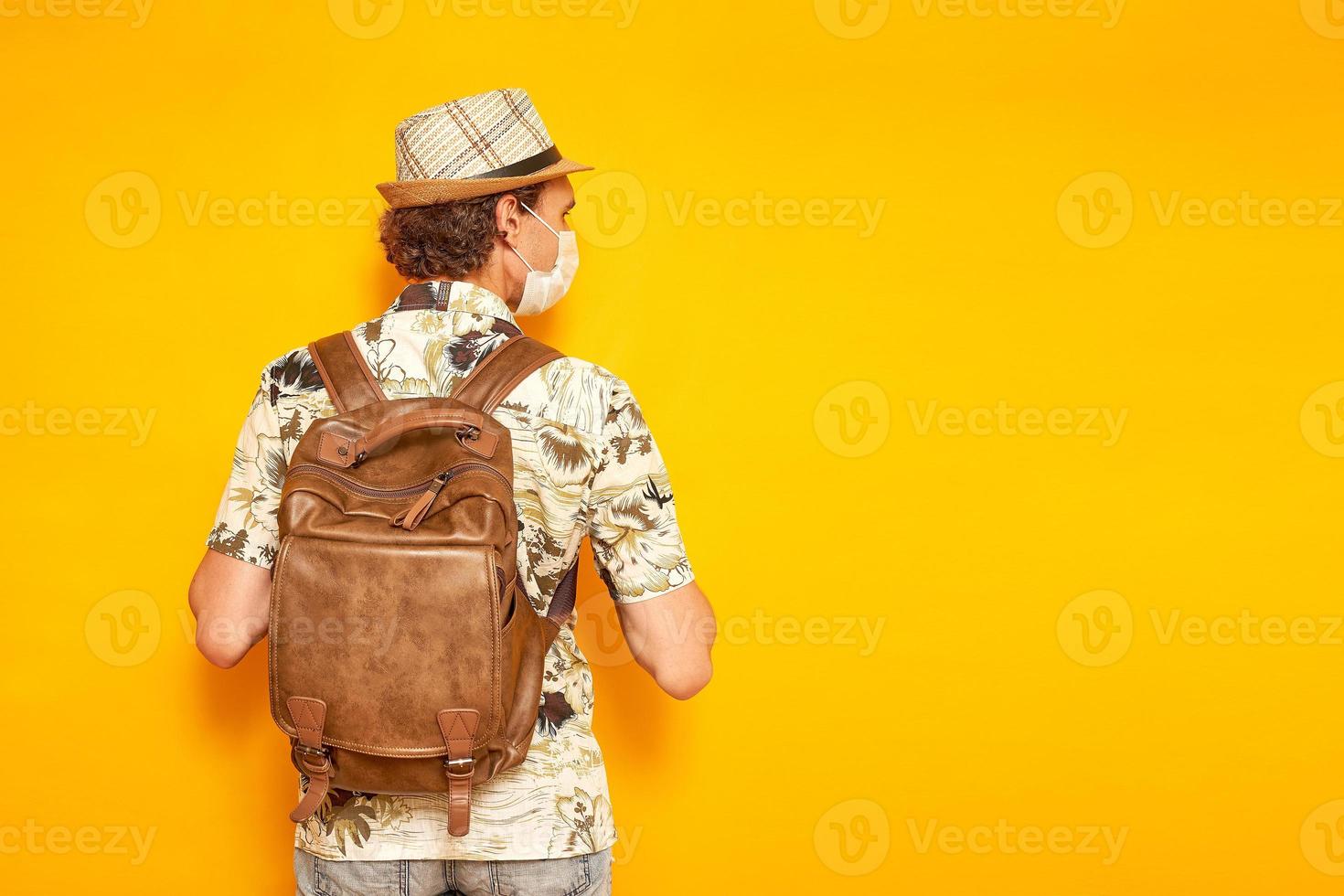 el viajero turista masculino con mochila con máscara médica se para con la espalda y mira a la distancia en el fondo. aislado en un fondo amarillo con espacio para texto. concepto de personas, viajes, vacaciones foto