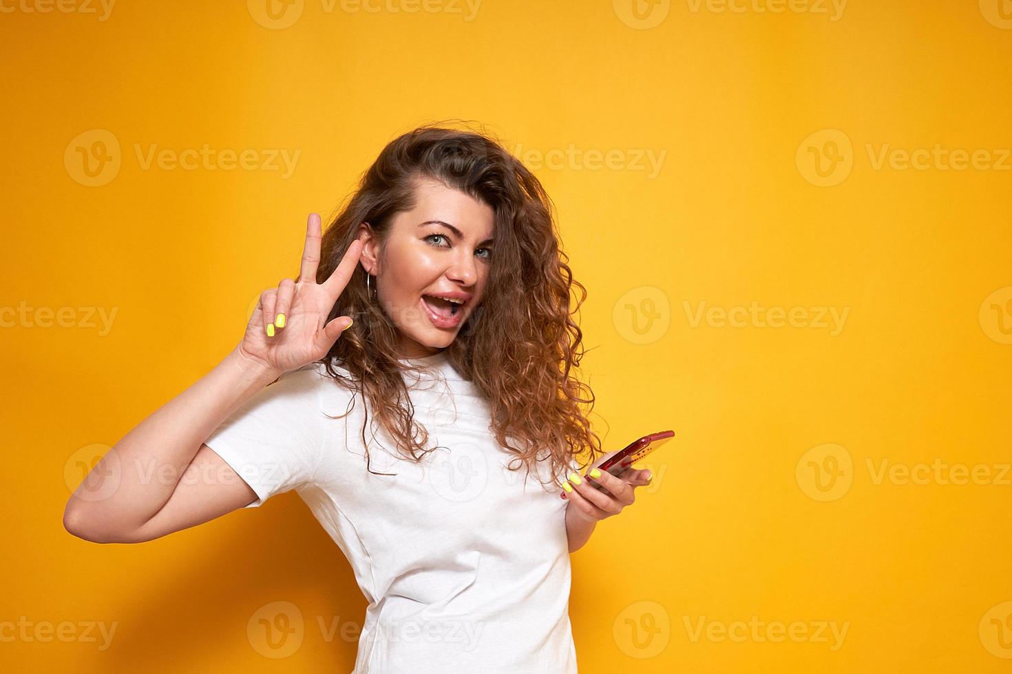 a happy smiling young woman in a white T-shirt and curly hair stands on a yellow isolated background with a phone in her hand and shows a victorious peaceful gesture. concept of people, technologies photo