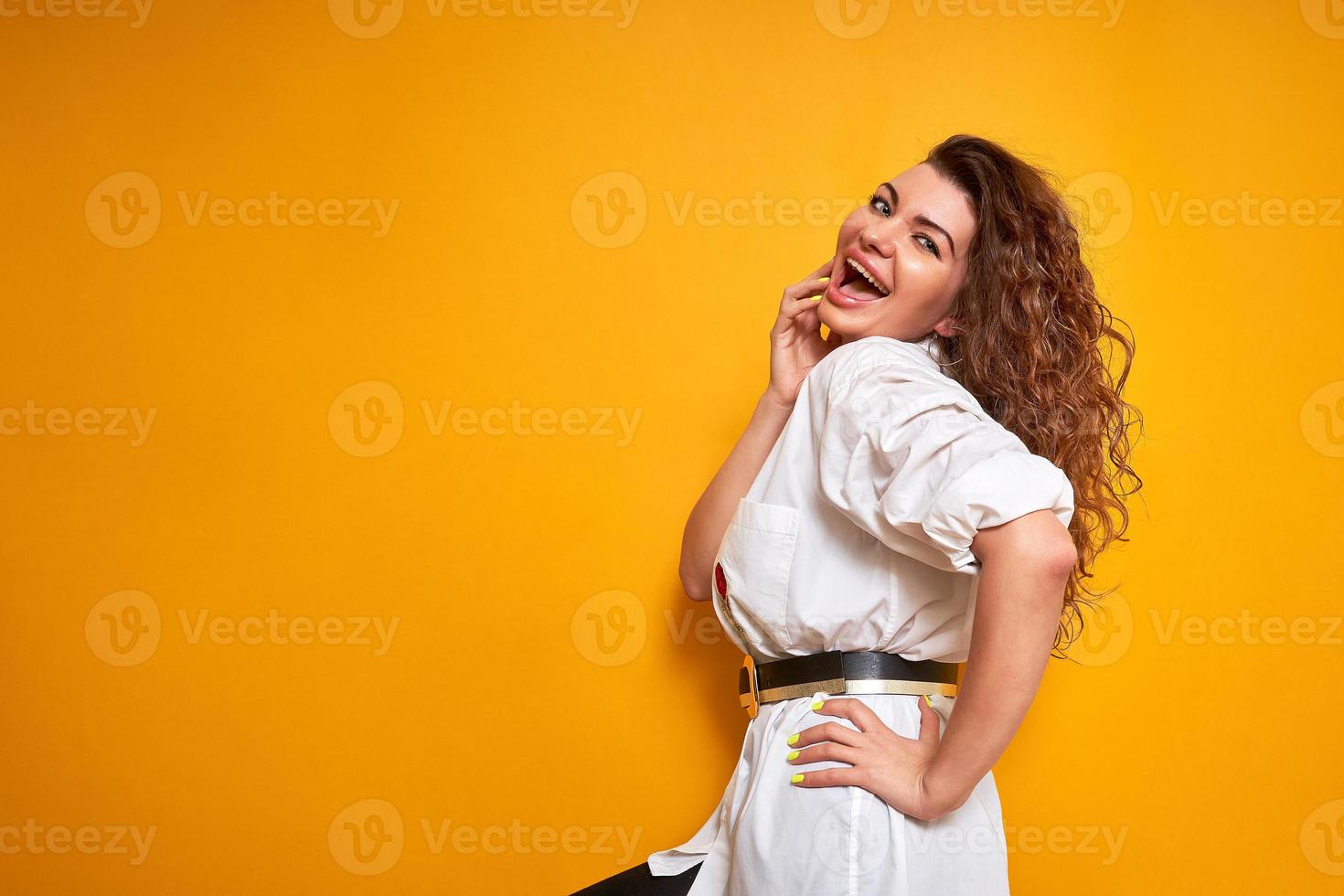 an excited joyful young woman, pleasantly surprised by good news about discounts, promotions, sales, holds her hand near her face, dressed in white shirt, stands sideways on yellow isolated background photo