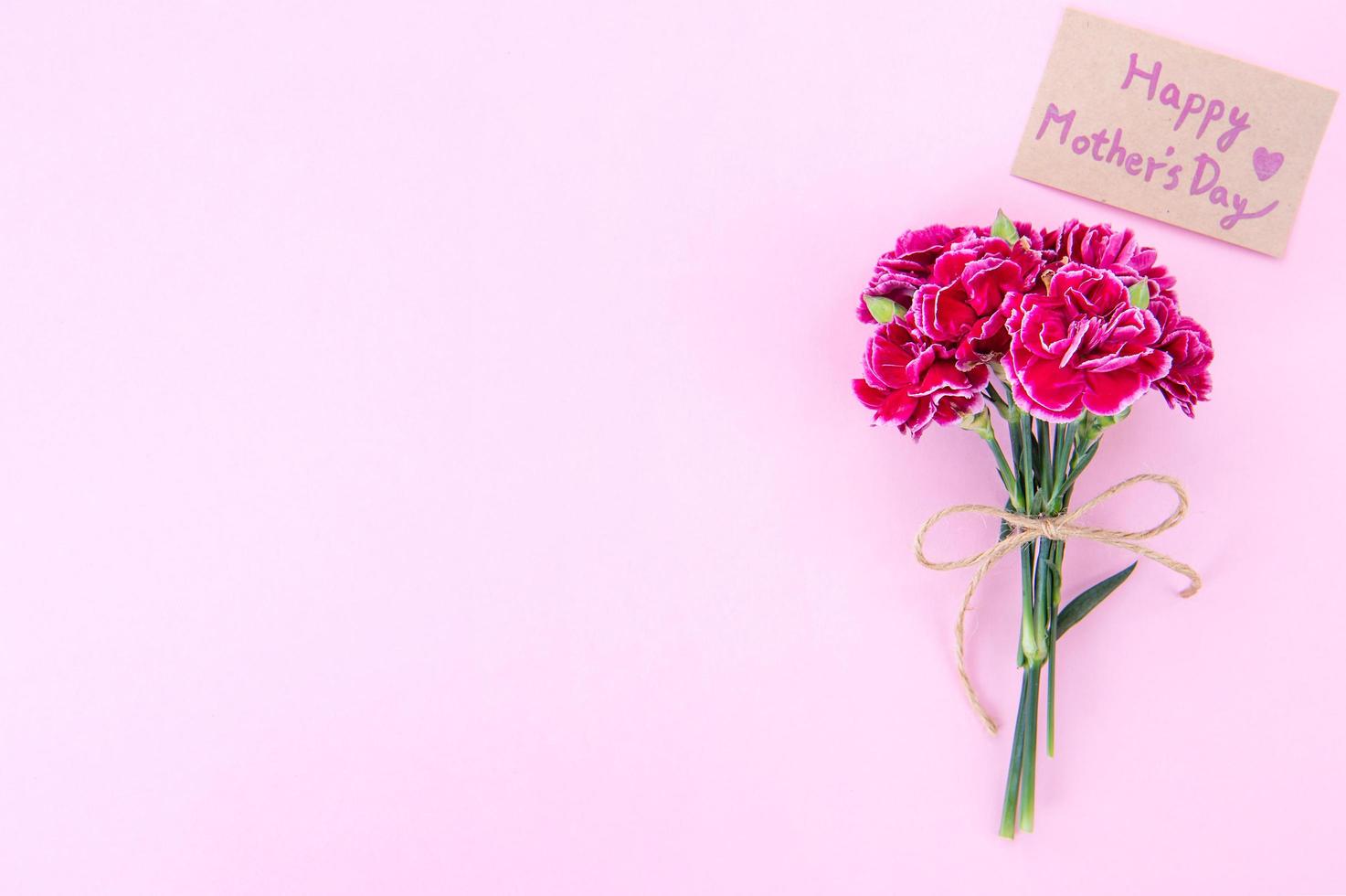 fotografía conceptual de la idea del día de las madres: hermosos claveles florecientes atados por un lazo con una tarjeta de texto kraft aislada en una mesa moderna y brillante, espacio de copia, capa plana, vista superior foto