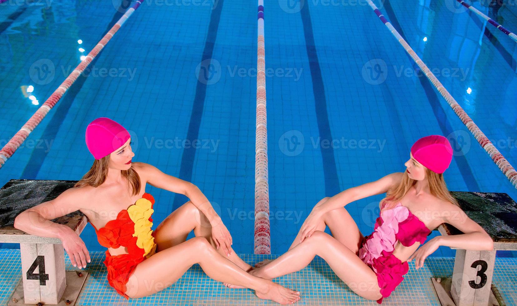 dos hermosas y asombrosas mujeres rubias delgadas jóvenes con estilo en trajes de baño coloridos y sombreros de natación rosa en la piscina. el verano. concepto de vacaciones y deporte. foto