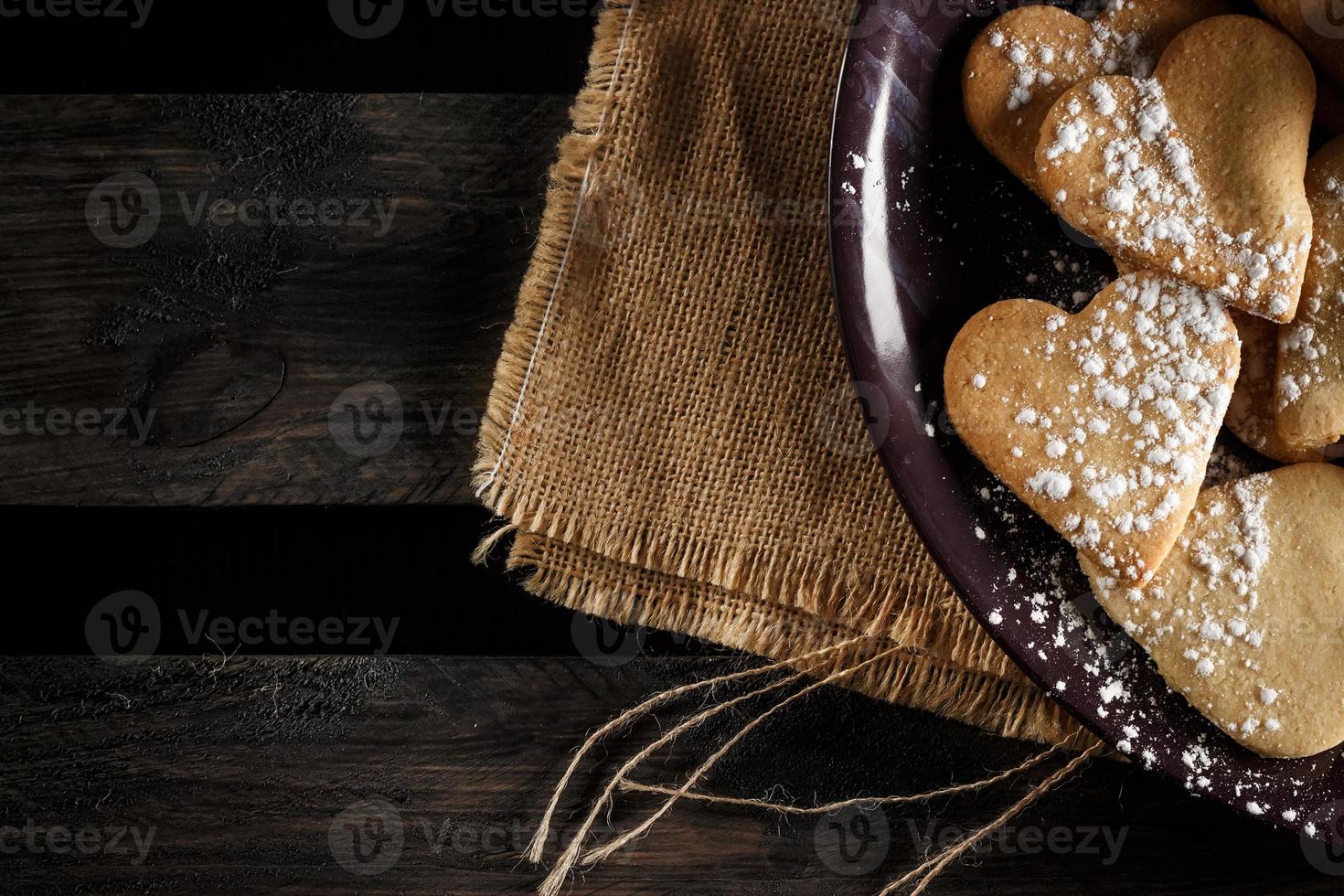 deliciosas galletas caseras en forma de corazón espolvoreadas con azúcar glaseado sobre tela de saco y tablas de madera. imagen horizontal vista desde arriba. foto