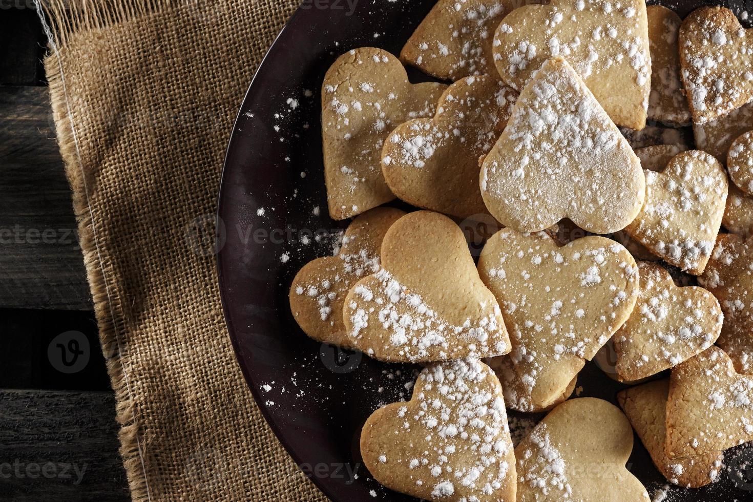 deliciosas galletas caseras en forma de corazón espolvoreadas con azúcar glaseado sobre tela de saco y tablas de madera. imagen horizontal vista desde arriba. foto