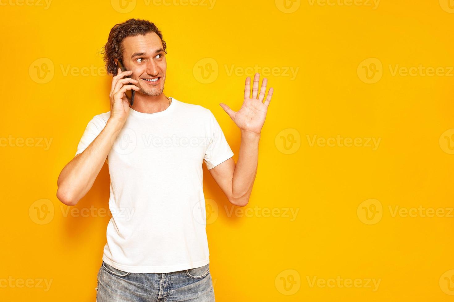 smiling man is talking on phone gesticulating with his raised hand, enjoying communication, dressed in casual clothes. isolated on studio yellow background. concept - people, technology, communication photo