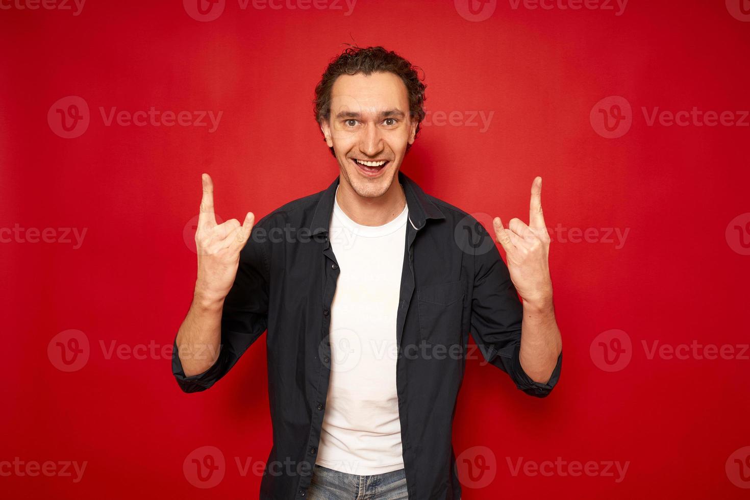 Happy overjoyed beautiful male with curly hair, shows cool sign love, says Yeah, that wonderful Pretty young man gestures indoor against red background copy space. People and body language concept photo